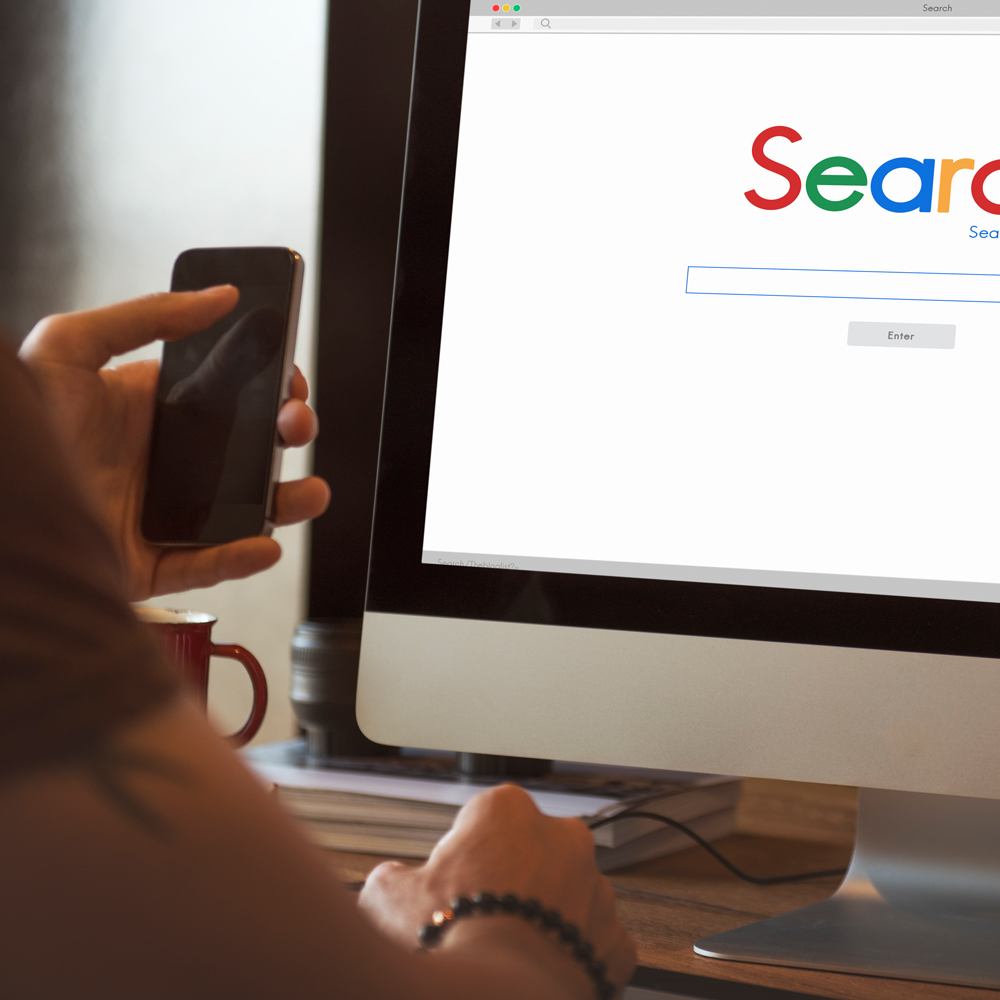 Person mit Smartphone vor Monitor mit Suchmaschine